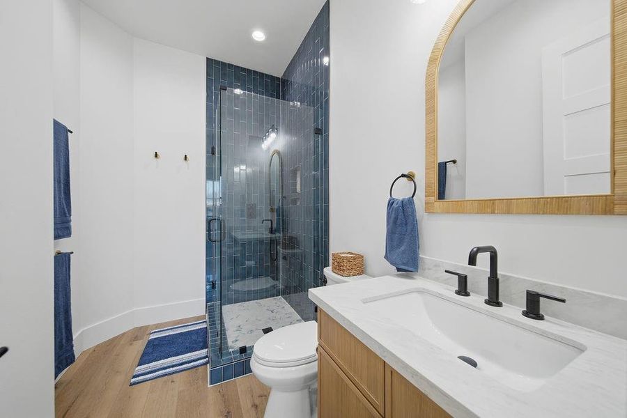 This modern bathroom features a sleek glass-enclosed shower with stylish blue tiles, a white vanity with dual black faucets, and warm wood accents. The space is enhanced by wood flooring, blue accessories, and ample lighting, creating a fresh and inviting atmosphere.
