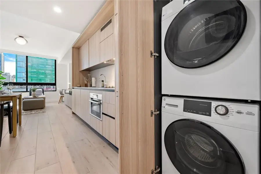 Built-in cabinetry featuring light wood-finish flat-panel doors, integrated stackable laundry appliances, integrated oven, and wood-finish flooring Built-in cabinetry featuring light wood-finish flat-panel doors, integrated stackable laundry appliances, integrated oven, and wood-finish flooring