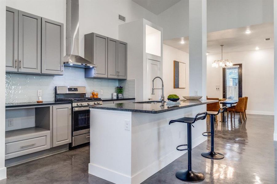 Kitchen with wall chimney range hood, an island with sink, stainless steel stove, sink, and gray cabinets