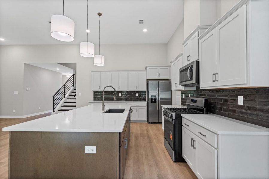 Modern kitchen featuring a large island with a sink, stainless steel appliances, extensive cabinetry, a dark subway tile backsplash, and wood-finish flooring Modern kitchen featuring a large island with a sink, stainless steel appliances, extensive cabinetry, a dark subway tile backsplash, and wood-finish flooring