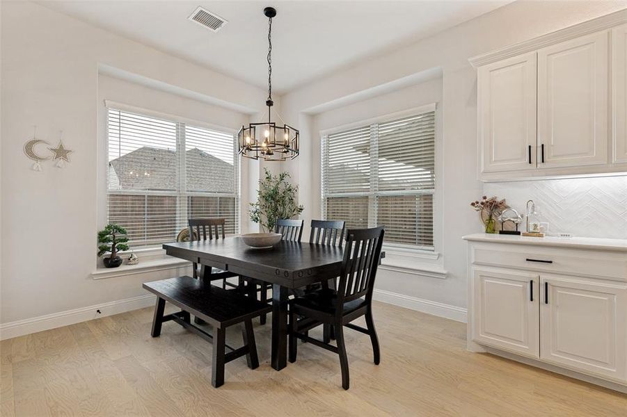 Bright dining area featuring light wood-finish flooring, a modern chandelier, and two large windows with blinds