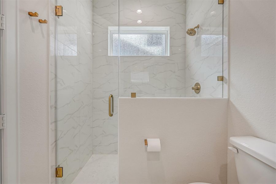 A frameless-glass walk-in shower completes the first-floor guest suite, featuring marble-look tile extending to the ceiling, a built-in niche, privacy window and warm brushed-gold fixtures.