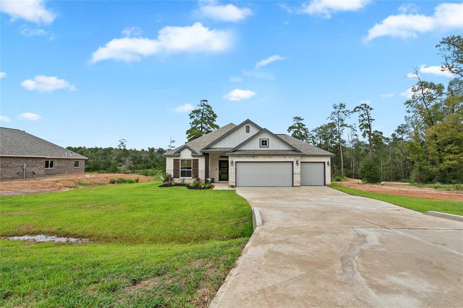 This is a newly constructed single-story home featuring a large front yard, an attached three-car garage, and a concrete driveway. The house is set in a developing neighborhood with natural surroundings.