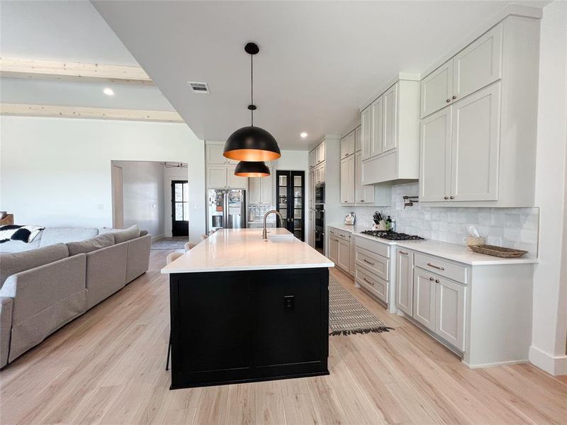 Kitchen with dark cabinets, decorative light fixtures, tasteful backsplash, an island with sink, and light wood-type flooring