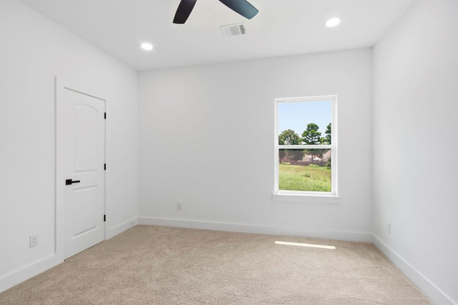 Bright and inviting room with plush carpeting, a modern ceiling fan, and a window offering serene views of greenery.