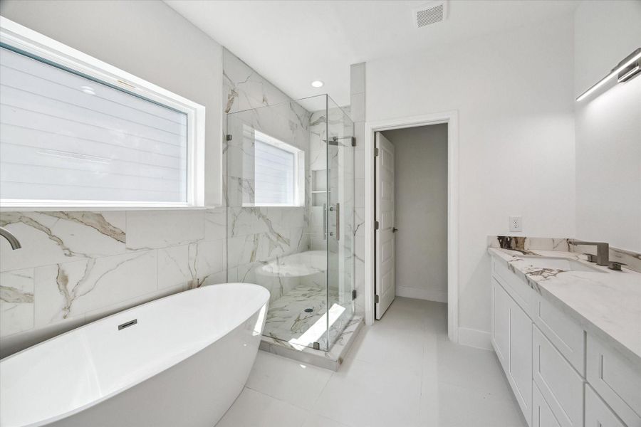 The primary bathroom delivers a spa-inspired atmosphere with a freestanding soaking tub, frameless glass shower, and striking marble-look tile carried across the walls and shower floor. Natural light streams through multiple windows, while the long vanity provides generous storage and counter space. Sleek fixtures and a bright neutral palette complete this polished, contemporary retreat.