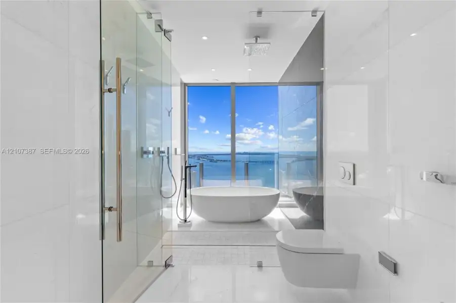 Freestanding soaking tub, floor-to-ceiling glass-enclosed shower, wall-mounted commode, large-format tile flooring, and floor-to-ceiling windows with coastal scenery