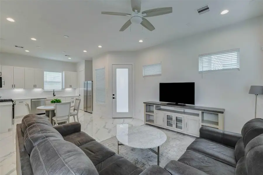 Open-concept living area with seamless flow into the dining space and kitchen. The room features glossy marble-look tile floors, recessed lighting, ceiling fan, window coverings, furnished seating, media console, and a bright, airy layout designed for comfortable daily living.