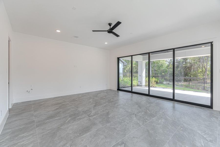 This spacious, modern living area features sleek gray tile flooring, a ceiling fan, and large sliding glass doors offering abundant natural light and views of the greenery outside.
