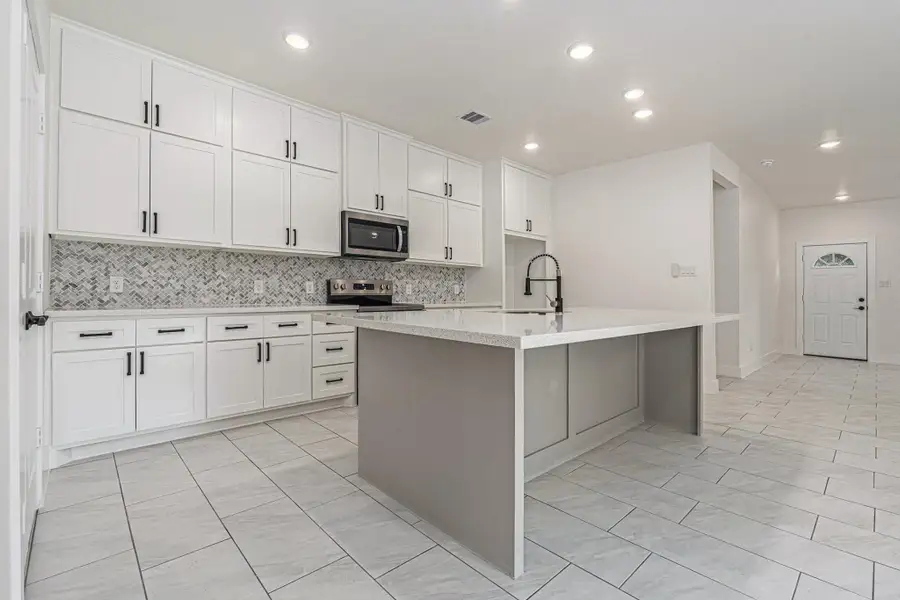 This photo showcases a modern kitchen with sleek white cabinetry, a stylish herringbone backsplash, and stainless steel appliances. It features a spacious island with a built-in sink and ample counter space, complemented by recessed lighting and a light tile floor for a bright and airy feel.