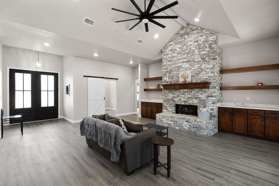 Living room featuring a barn door, beamed ceiling, light wood-style flooring, a ceiling fan, and a brick fireplace