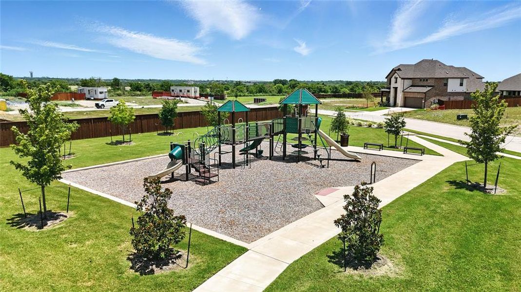Community playground featuring multiple slides and climbing structures, surrounded by a manicured lawn with mature trees and a concrete pathway