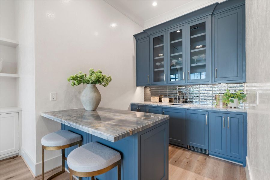 The wet bar off of the living room. Vibrant quartzite countertops and mirrored tile backsplash contrast the deep blue-gray of the woodwork. Shelves and cabinets are ideal for displaying glassware and more. The space includes a clear-ice maker and a beverage chiller. Add in some bar stools to tie everything together!