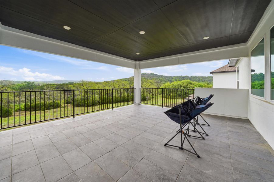 This expansive covered patio features a tiled floor, black metal railings, and recessed lighting, offering elevated views of the green landscape
