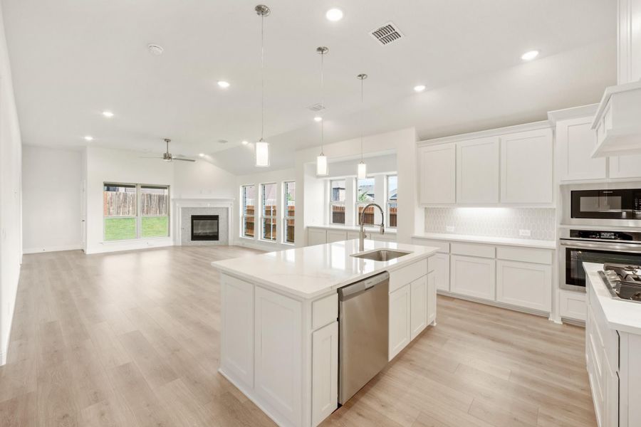 Open-concept kitchen with white cabinets, large island, stainless steel appliances, and light wood floors opening to living room with fireplace.