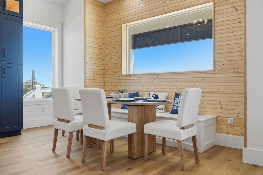 This cozy dining nook features a wood-paneled accent wall, built-in bench seating with blue and white cushions, a modern wood table, and upholstered chairs. Large windows provide ample natural light and a view outside. The space offers a blend of comfort and style, perfect for casual dining.