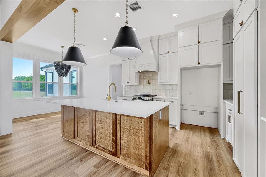 Kitchen featuring tasteful backsplash, light wood-type flooring, premium range hood, a kitchen island with sink, and hanging light fixtures