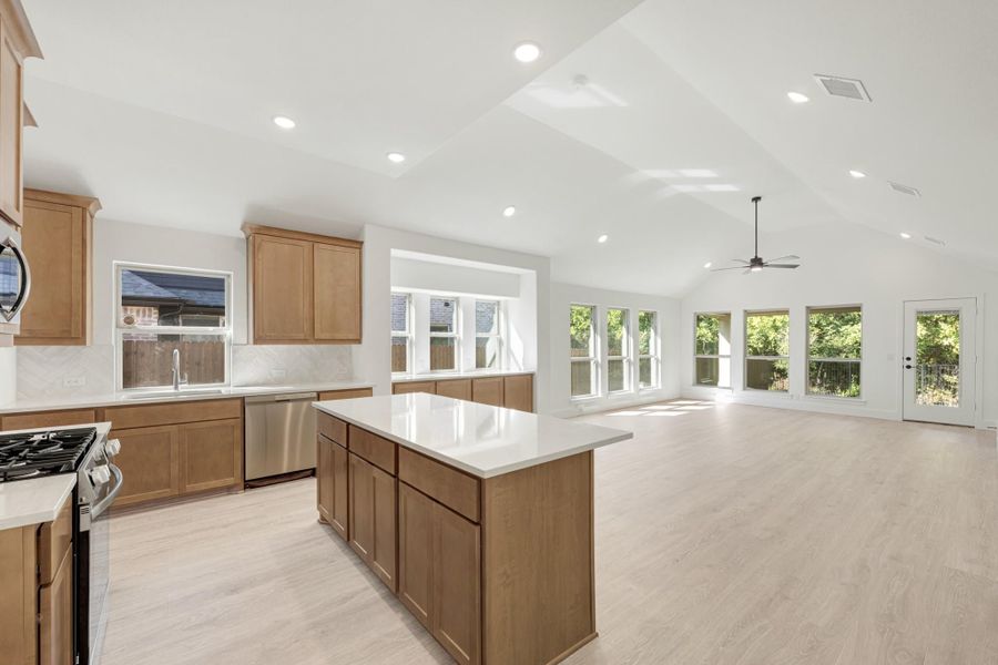 Open kitchen with wood cabinets, center island, and vaulted ceiling flowing into a bright living area with large windows