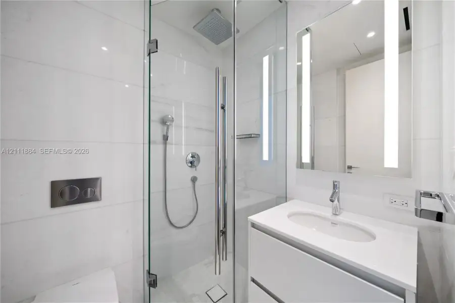 Bathroom featuring a frameless glass shower enclosure, oversized rain showerhead, wall-mounted dual-flush toilet, vanity with integrated sink, and vertical LED-integrated mirror Bathroom featuring a frameless glass shower enclosure, oversized rain showerhead, wall-mounted dual-flush toilet, vanity with integrated sink, and vertical LED-integrated mirror