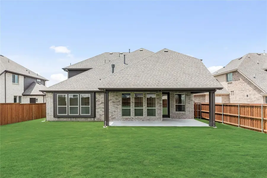 Rear view of brick home with covered patio, large windows, and fenced backyard with green lawn
