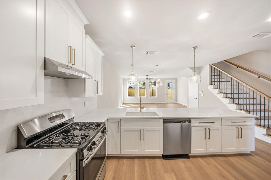 This modern kitchen features sleek white cabinetry, a stainless steel stove and dishwasher, and a spacious island. It opens to a bright living area with wood flooring and stylish pendant lighting, complemented by a staircase with a contemporary railing.
