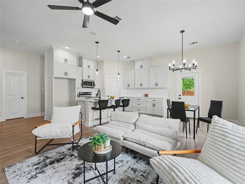 Open-concept living area featuring wood-finish flooring, white cabinetry, a kitchen island with bar seating, a dining area, and modern light fixtures Open-concept living area featuring wood-finish flooring, white cabinetry, a kitchen island with bar seating, a dining area, and modern light fixtures