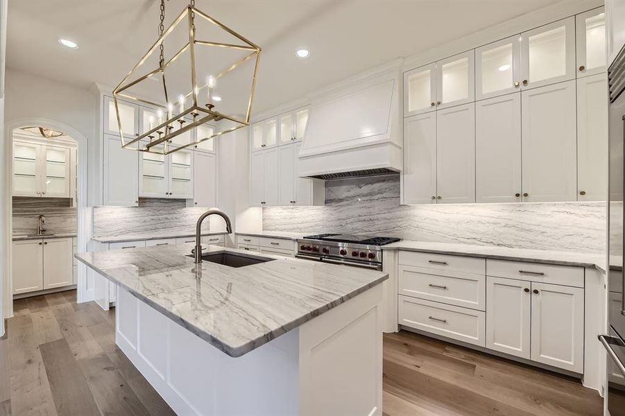 Kitchen featuring light stone counters, custom range hood, and cabinets extending to the ceiling with lighted glass cabinets