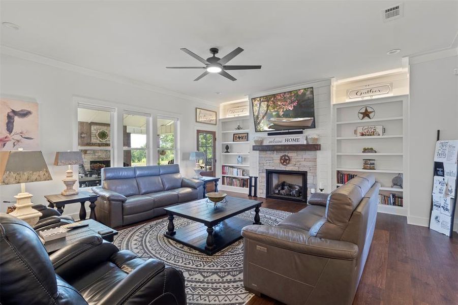 Inviting living room featuring custom built-ins, a stone fireplace with wood mantel, hardwood floors, and abundant natural light, creating a warm and comfortable gathering space.