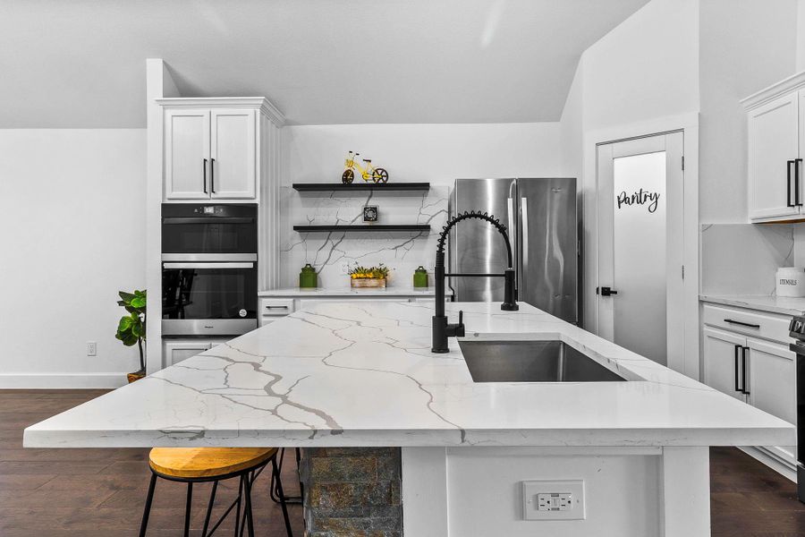 This modern kitchen features sleek white cabinetry, a spacious island with a built-in sink and black fixtures, stainless steel appliances, and a convenient pantry door. The open shelving and stylish decor add a contemporary touch.