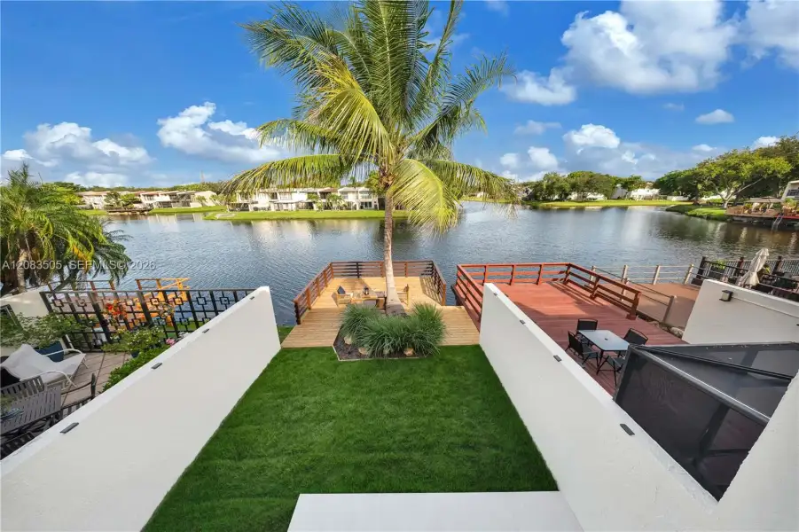Waterfront property featuring a private wooden deck, manicured lawn, perimeter privacy walls, and tropical landscaping Waterfront property featuring a private wooden deck, manicured lawn, perimeter privacy walls, and tropical landscaping
