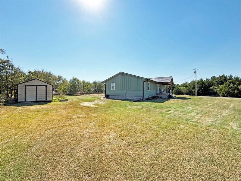 View of grassy yard featuring a storage shed, a porch, and view of scattered trees View of grassy yard featuring a storage shed, a porch, and view of scattered trees