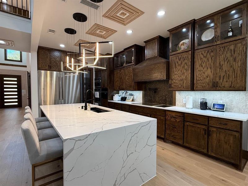 Kitchen featuring glass insert cabinets, hanging light fixtures, dark brown cabinetry, light stone countertops, and light wood finished floors