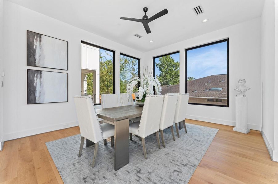 This dining room features a modern, spacious design with large windows offering abundant natural light. It includes a sleek wooden table with eight upholstered chairs, a stylish ceiling fan, and contemporary artwork. The room has light hardwood floors and a neutral color palette, creating an elegant atmosphere.