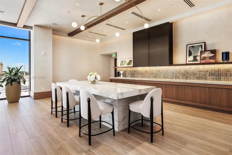 Waterfall-edge stone island with bar seating, custom wood-finish cabinetry, linear tile backsplash, wood-finish flooring, and recessed ceiling beams Waterfall-edge stone island with bar seating, custom wood-finish cabinetry, linear tile backsplash, wood-finish flooring, and recessed ceiling beams