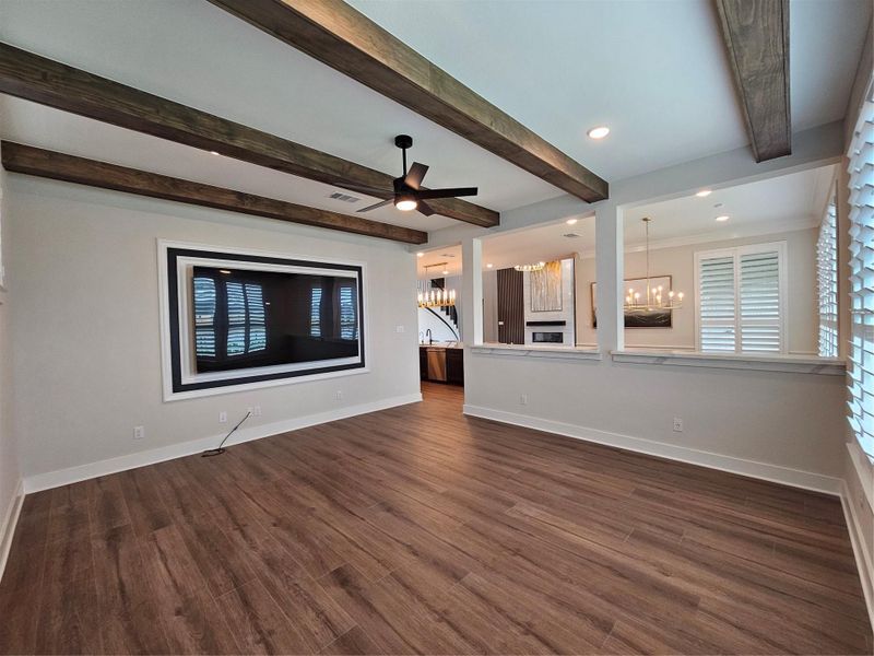 This spacious room features elegant wood flooring and exposed ceiling beams, offering a modern, open-concept layout. A stylish ceiling fan and recessed lighting enhance the ambiance, while large windows provide ample natural light. The room seamlessly connects to a kitchen area with a contemporary chandelier, ideal for entertaining.