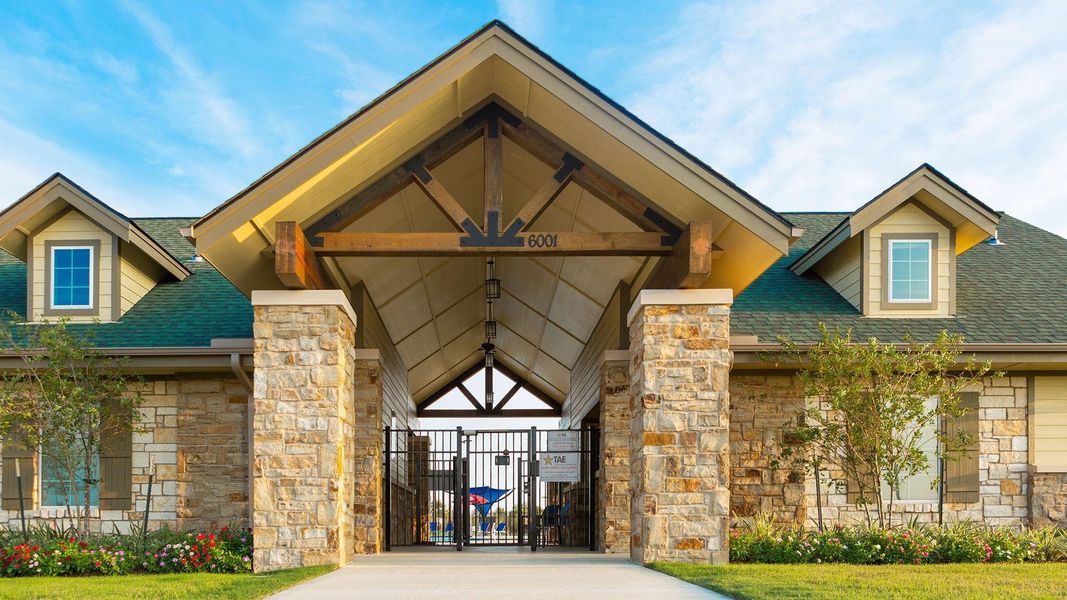 This photo features a welcoming entrance to a residential community, showcasing a rustic stone facade with a peaked roof and gated entry. The landscape is well-maintained, enhancing the inviting atmosphere.