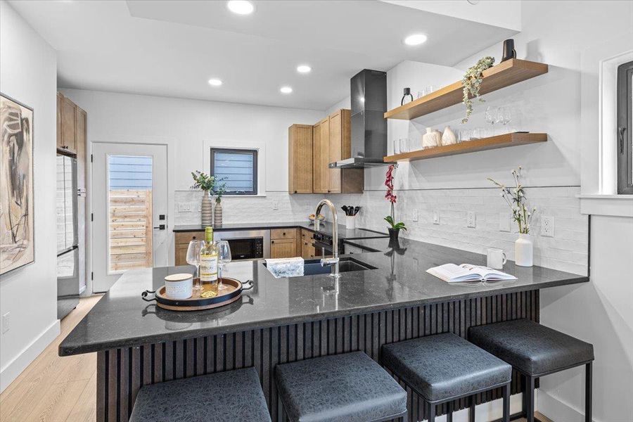 Kitchen with tasteful backsplash, light wood finished floors, dark stone counters, a peninsula, and wall chimney exhaust hood