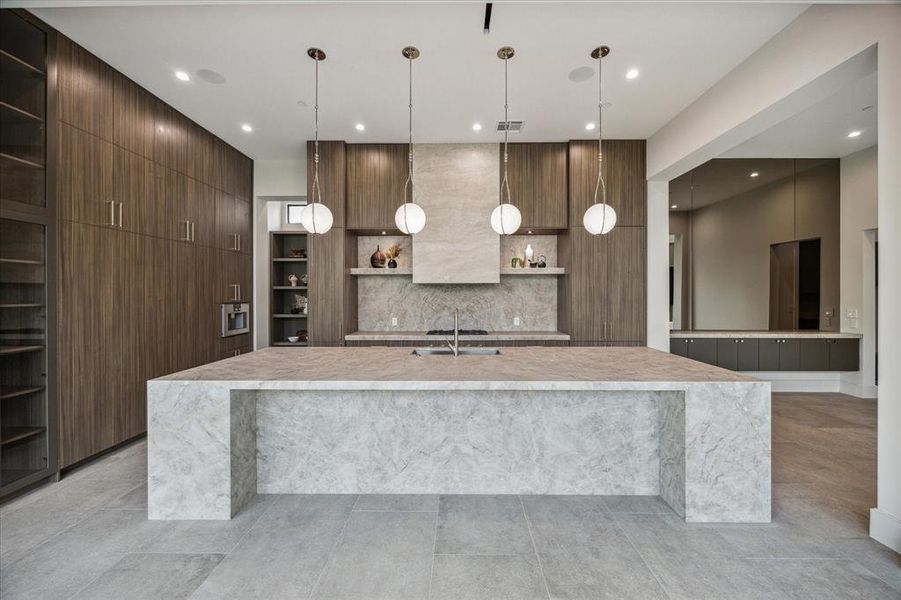 This luxurious kitchen focal point is a breakfast island with Taj Mahal Quartzite countertops and pull-up counter height seating, complemented by a stunning Taj Mahal wrapped vent hood and floating shelves. The expansive wall of steel and glass windows offers breathtaking viewsof the rear lawn and sparkling pool, flooding the space with natural light.