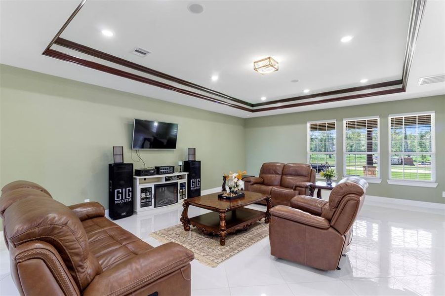 This spacious living room features sleek tile flooring, a coffered ceiling with recessed lighting, and large windows offering plenty of natural light. It includes comfortable leather seating, a modern entertainment setup with a wall-mounted TV, and a cozy atmosphere perfect for relaxation or entertaining.