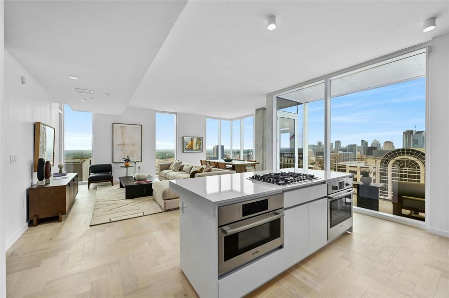 Open-concept living area featuring light wood-finish flooring, a contemporary kitchen island with a gas cooktop and built-in oven, and a wall of floor-to-ceiling windows