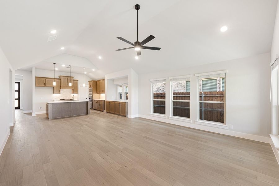 Open living area with vaulted ceiling, ceiling fan, hardwood floors, and open kitchen with wood cabinets and island in background Open living area with vaulted ceiling, ceiling fan, hardwood floors, and open kitchen with wood cabinets and island in background