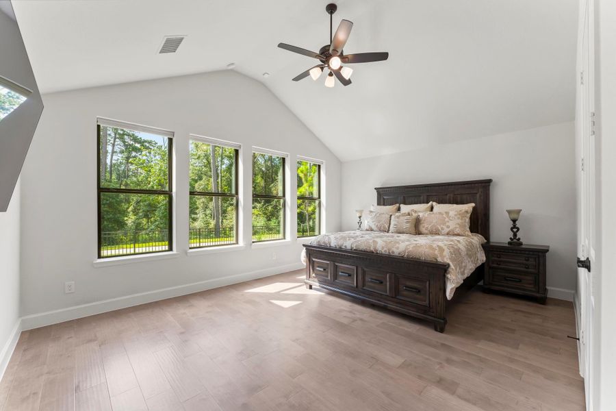 Spacious primary bedroom with vaulted ceilings and light wood floors. Three tall windows with dark frames offer a view of the outdoors. The room is painted white, and a ceiling fan is mounted in the center. Spacious primary bedroom with vaulted ceilings and light wood floors. Three tall windows with dark frames offer a view of the outdoors. The room is painted white, and a ceiling fan is mounted in the center.
