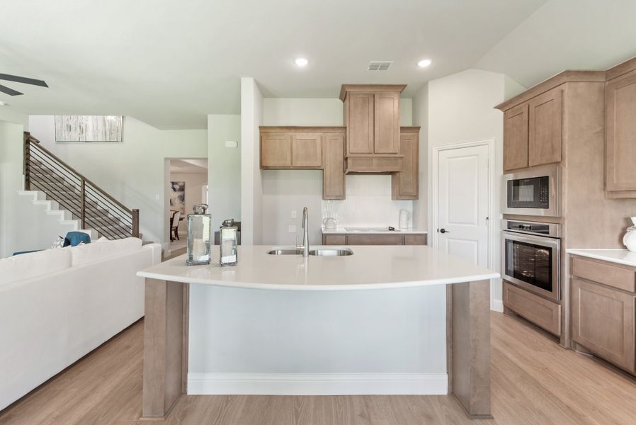 Open kitchen with curved island, tan wood cabinets, stainless steel appliances, and light hardwood floors