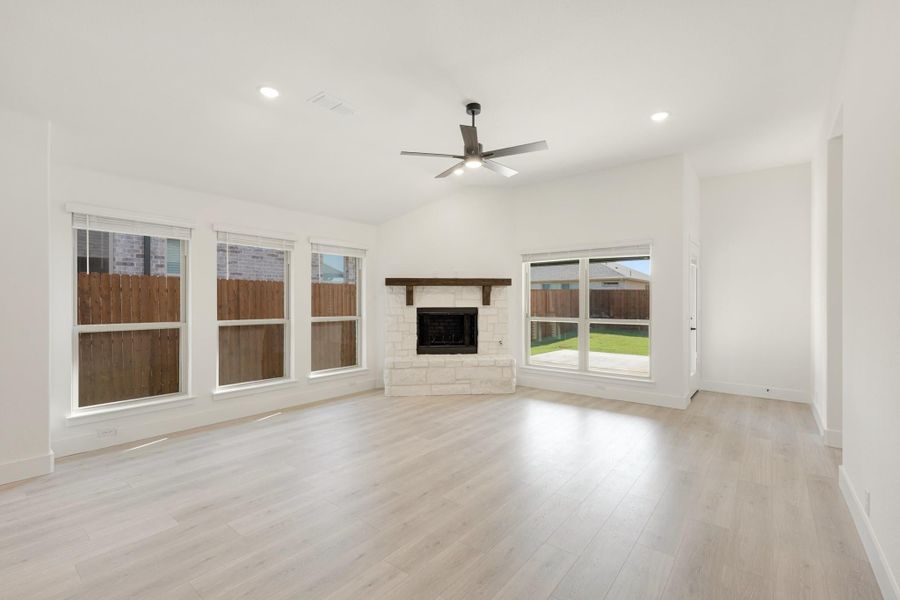Open living room with stone fireplace, ceiling fan, light hardwood floors, and large windows overlooking backyard Open living room with stone fireplace, ceiling fan, light hardwood floors, and large windows overlooking backyard