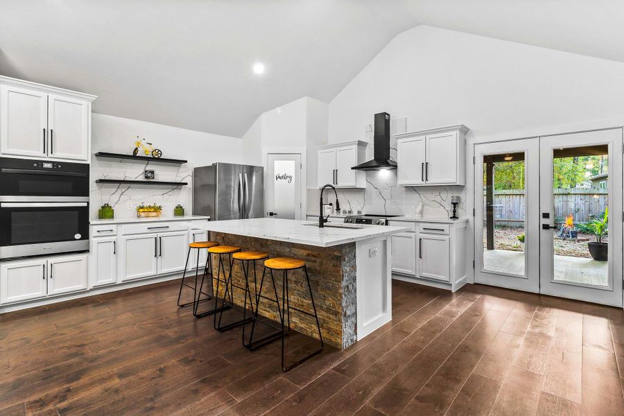 This modern kitchen features high ceilings, sleek white cabinetry, and stainless steel appliances. It boasts a large center island with seating and a stone accent, wood flooring, and French doors leading to a backyard, providing a bright and inviting space. This modern kitchen features high ceilings, sleek white cabinetry, and stainless steel appliances. It boasts a large center island with seating and a stone accent, wood flooring, and French doors leading to a backyard, providing a bright and inviting space.