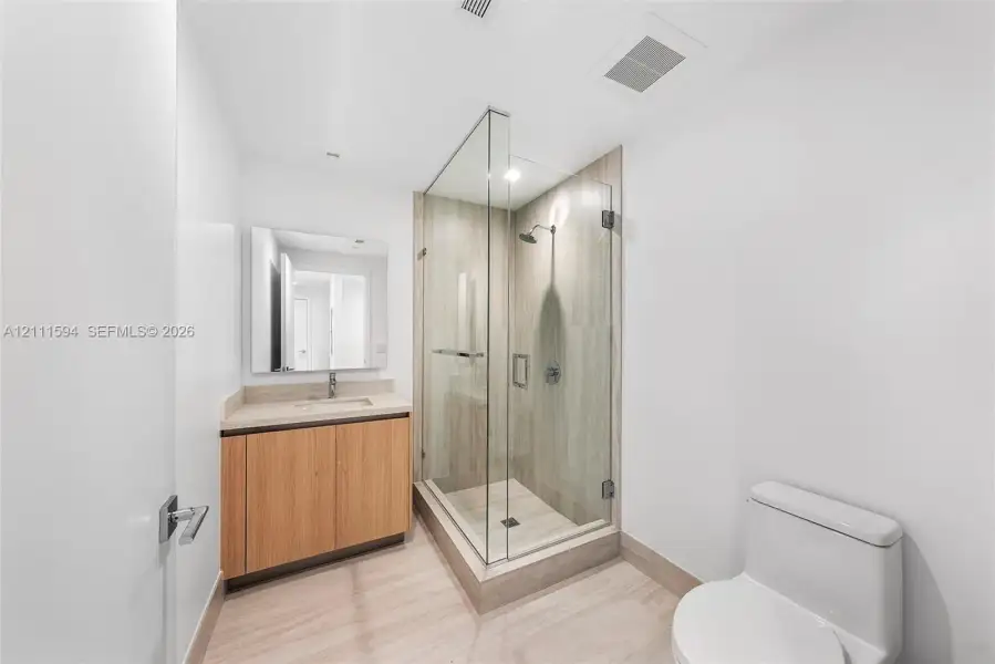 Walk-in shower with frameless glass enclosure and tile surround, wood-finish cabinetry, rectangular under-mount sink, and neutral tile flooring Walk-in shower with frameless glass enclosure and tile surround, wood-finish cabinetry, rectangular under-mount sink, and neutral tile flooring