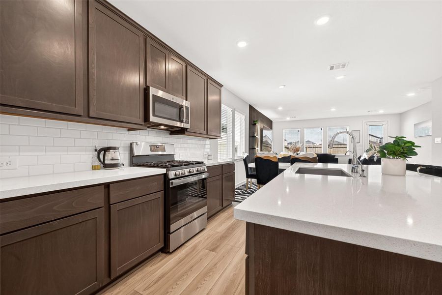 Open-concept kitchen and living area featuring a large quartz island with seating, rich cabinetry, stainless steel appliances, subway tile backsplash, and seamless flow into the main living space. The thoughtfully designed layout is ideal for entertaining while offering a warm, modern aesthetic with abundant natural light throughout.