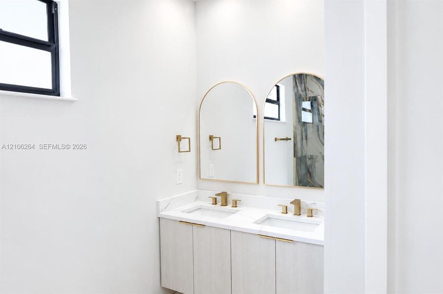 Dual-sink vanity featuring light wood-finish cabinetry, white stone-surfaced countertops, brushed gold-tone faucets, and twin arched mirrors