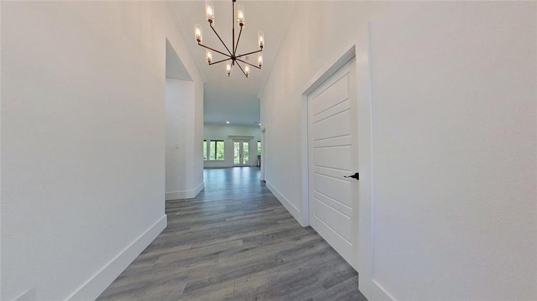 Hallway featuring light gray wood-finish flooring, white baseboards, and a contemporary linear chandelier with multiple light sources Hallway featuring light gray wood-finish flooring, white baseboards, and a contemporary linear chandelier with multiple light sources