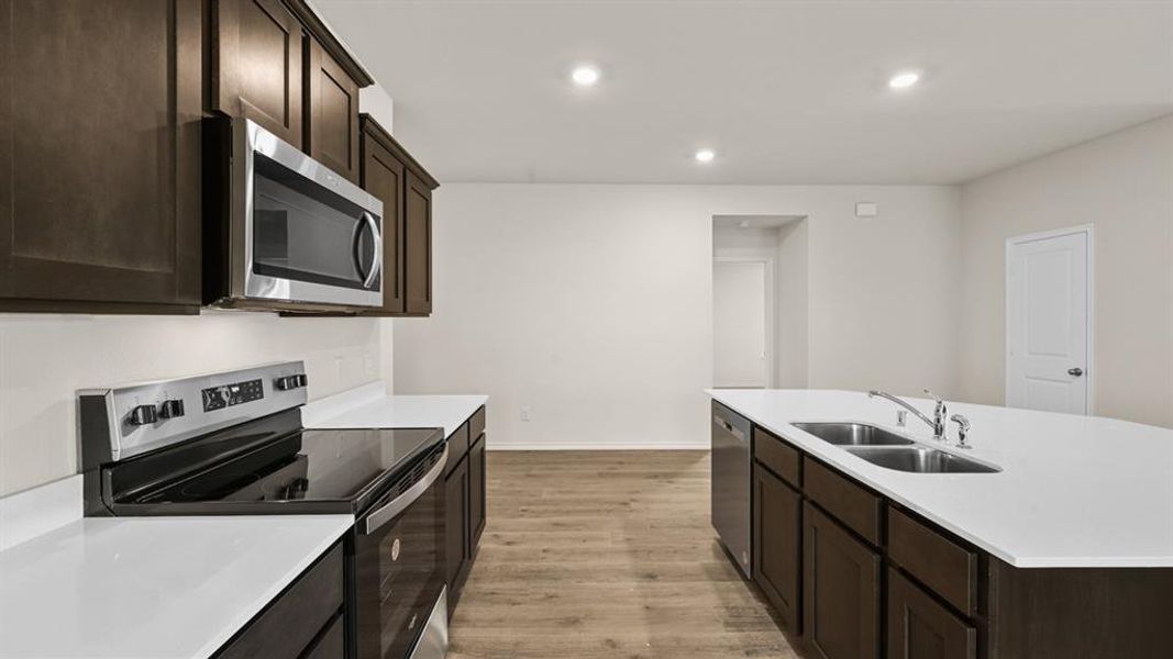 Kitchen featuring wood-finish flooring, dark wood cabinetry, white countertops, stainless steel appliances, and an island with a dual-basin sink Kitchen featuring wood-finish flooring, dark wood cabinetry, white countertops, stainless steel appliances, and an island with a dual-basin sink